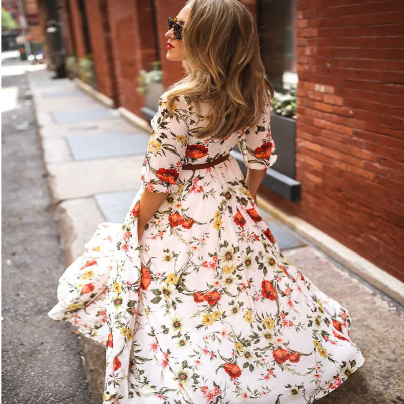 robe blanche à imprimé floral rouge et jaune, manches 3/4, taille marquée par une ceinture, jupe longue fluide et évasée en mouvement dans une rue.