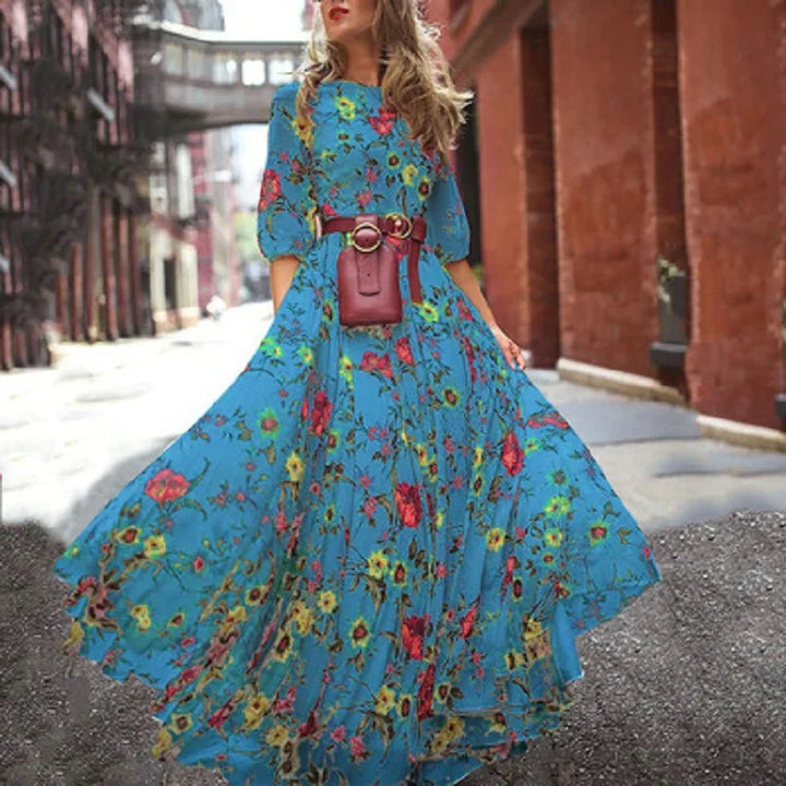 Femme portant la robe maxi longue  Estelle robe bleue à imprimé floral multicolore, manches 3/4, taille ceinturée avec sac ceinture, jupe longue évasée et fluide en mouvement dans une rue urbaine.