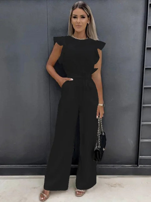 Femme debout portant une combinaison noire moderne à coupe large, sans manches, avec taille cintrée et pantalon fluide, offrant une silhouette élégante et contemporaine, accessoirisée d’un petit sac à main et de sandales à talons.