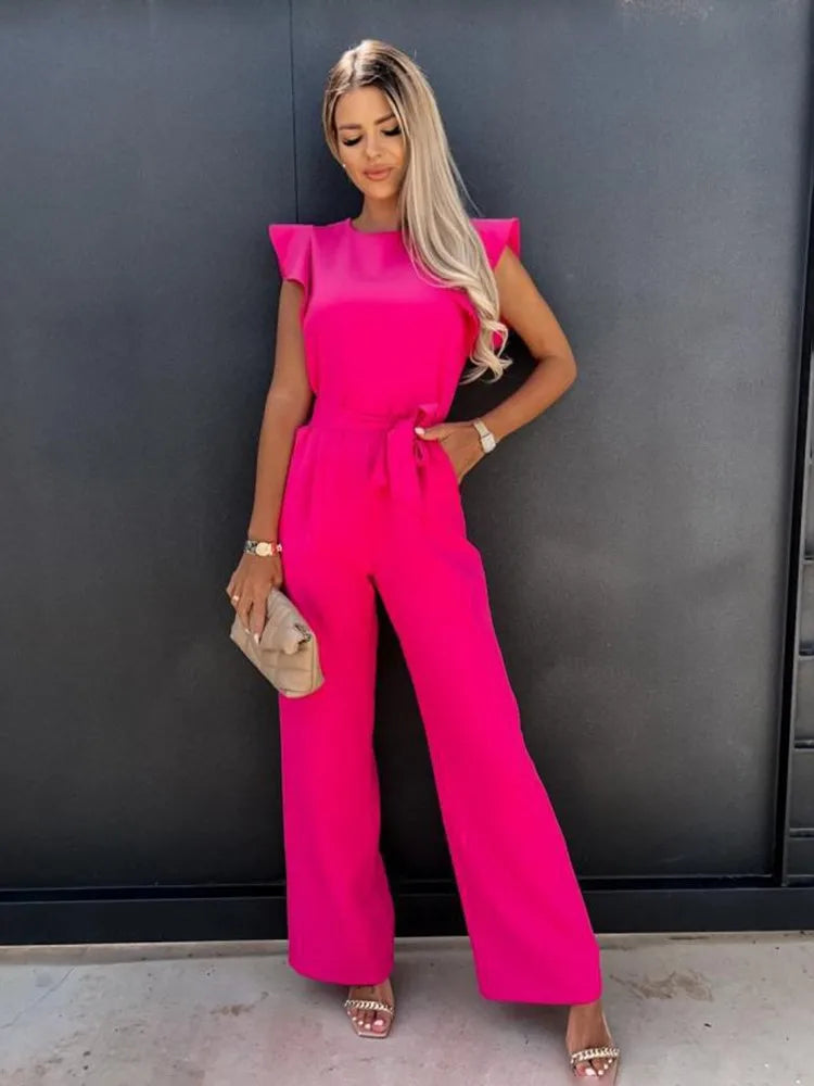 Femme debout portant une combinaison large rose fuchsia au style moderne, sans manches, avec taille ajustée et jambes fluides, créant une allure élégante et féminine, complétée par une pochette claire et des sandales à talons.