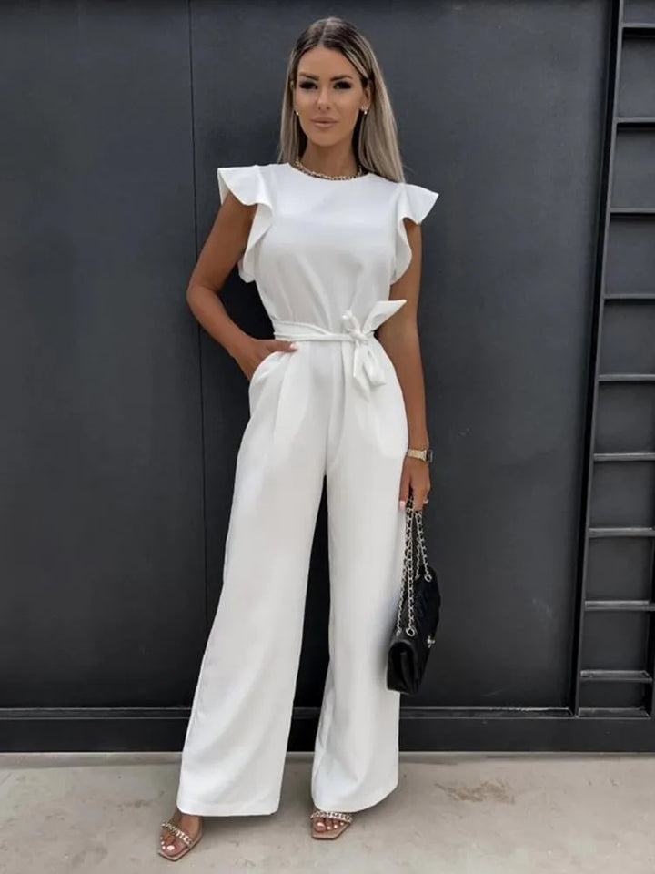Femme debout portant une combinaison blanche moderne à coupe large, sans manches, avec épaules structurées, taille nouée et pantalon fluide, offrant une allure élégante et raffinée, accessoirisée d’un sac à main noir et de sandales à talons.