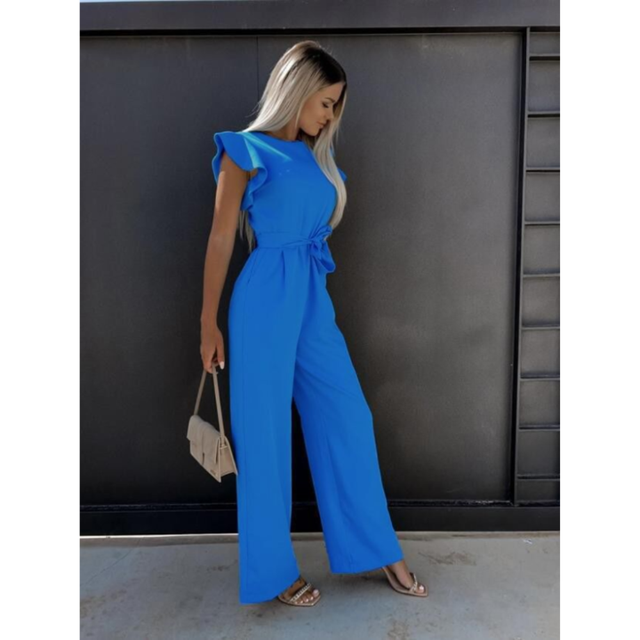 Femme de profil portant une combinaison large bleue au style moderne, sans manches avec épaules légèrement volantées, taille nouée et pantalon fluide, offrant une silhouette élégante et contemporaine, accompagnée d’un petit sac clair et de sandales plates.