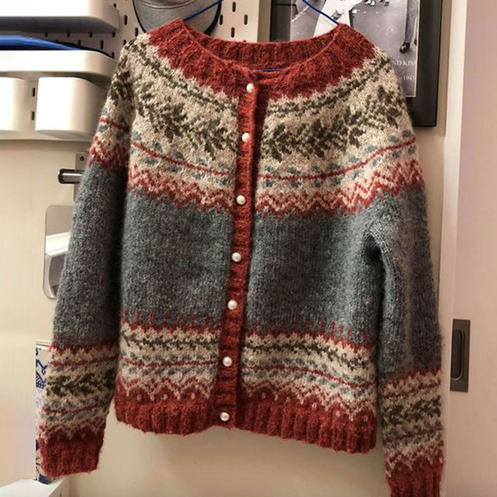 Cardigan pour femme en maille douce, présentant un motif nordique traditionnel dans des tons gris, rouge brique et beige, avec une fermeture à boutons nacrés et une coupe confortable suspendue sur un cintre.