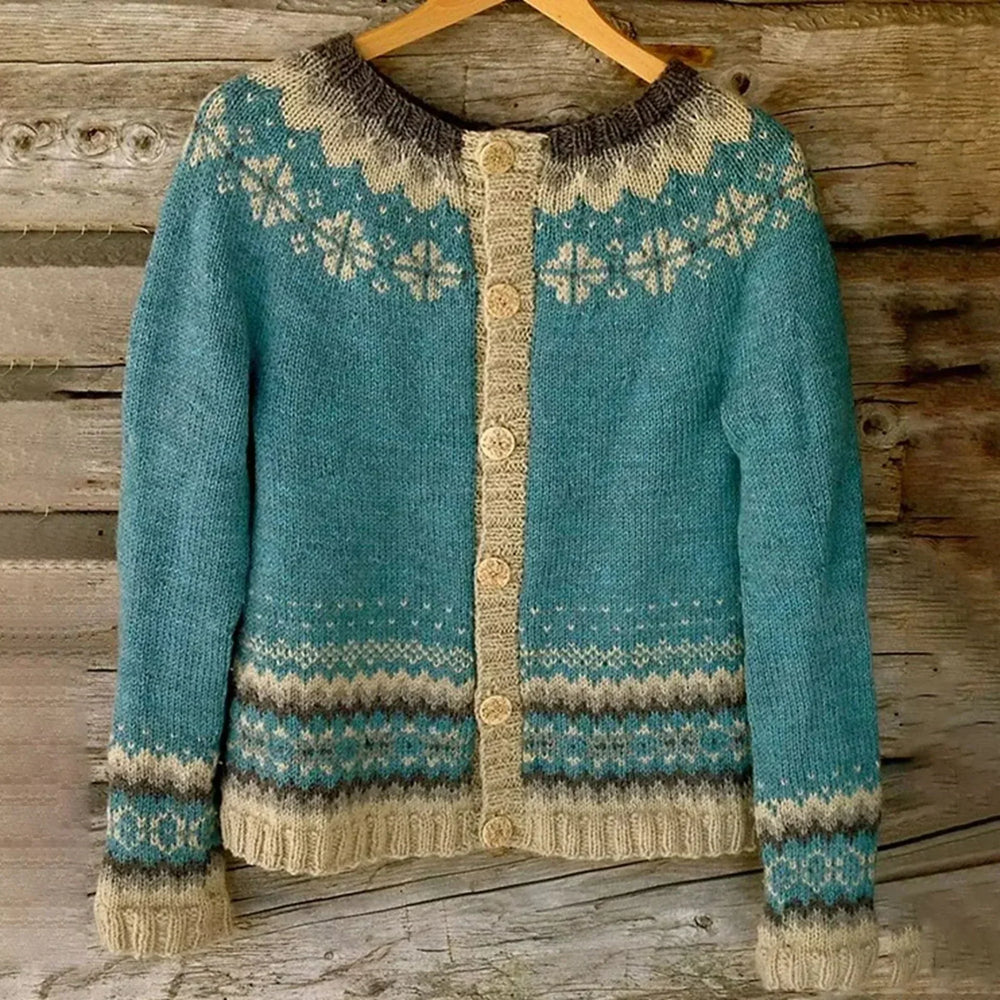 Cardigan pour femme en maille douce, de couleur bleu turquoise, orné de motifs nordiques beige et gris sur le col, les poignets et le bas, avec une rangée de boutons tricotés assortis, suspendu à un cintre devant un fond en bois rustique.