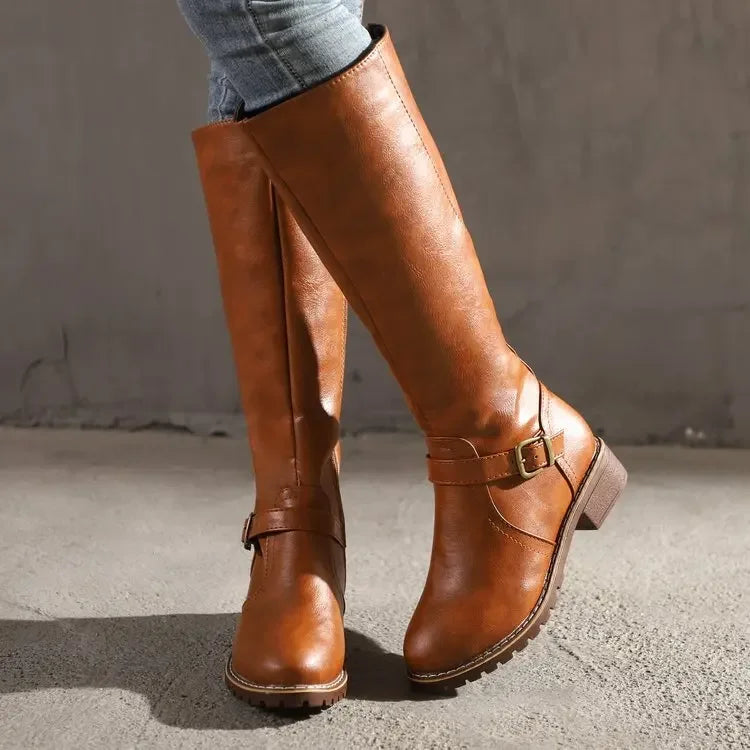 Bottes hautes en cuir synthétique marron pour femme, avec une boucle décorative dorée et une semelle antidérapante. Design élégant et confortable, parfait pour la saison hivernale. Portées avec un jean ajusté, elles offrent un style moderne et chaleureux, idéal pour les journées froides.