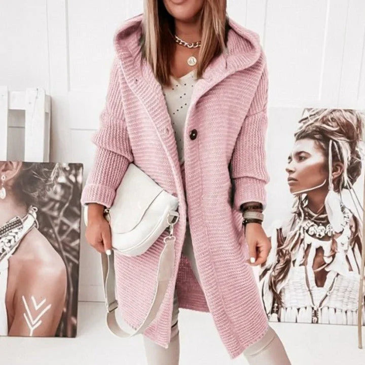 Femme portant un long cardigan en maille rose avec capuche et manches longues, combiné à un sac blanc et un pantalon clair, posant dans une pièce décorée de portraits artistiques.