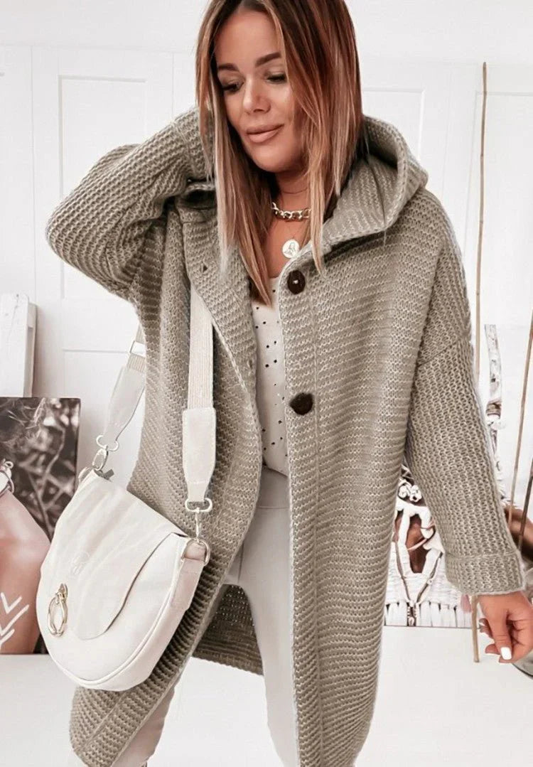 Femme portant un long cardigan kaki en grosse maille à capuche et gros boutons, assorti à un sac blanc et un pantalon clair, posant dans une pièce décorée de portraits artistiques.