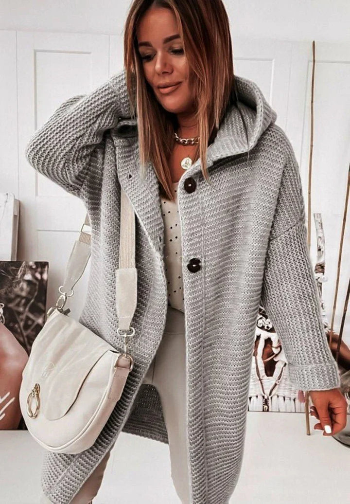 Femme portant un long cardigan en maille épaisse gris clair avec capuche et gros boutons, assorti à un sac blanc et un pantalon clair, posant dans une pièce décorée de portraits artistiques.