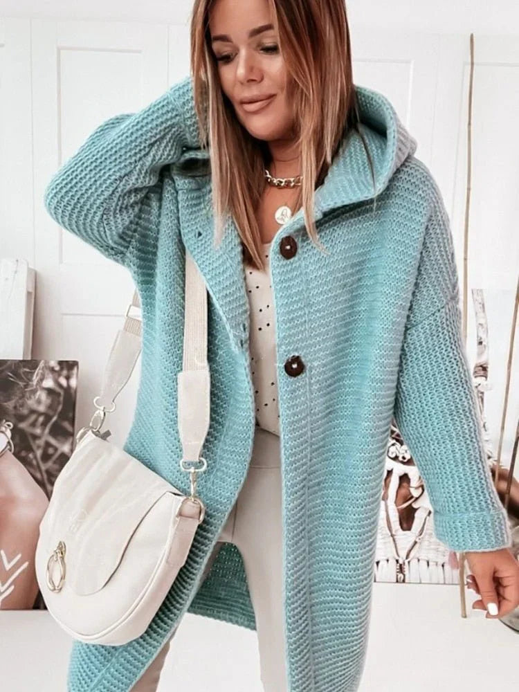 Femme portant un long cardigan vert en grosse maille à capuche et gros boutons, assorti à un sac blanc et un pantalon clair, posant dans une pièce lumineuse décorée de portraits artistiques.
