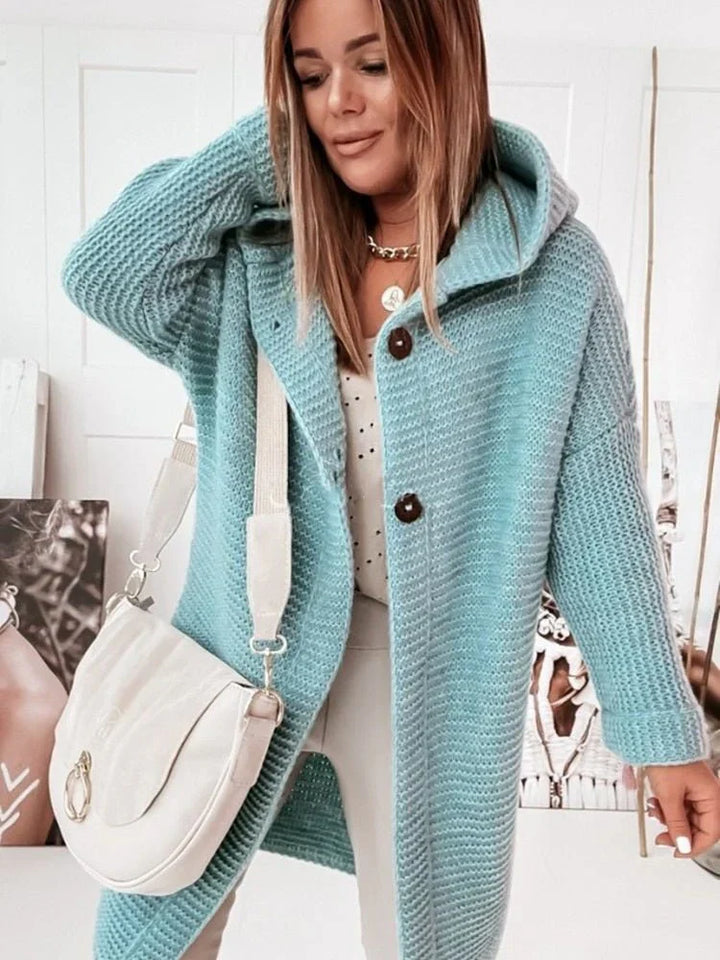 Femme portant un long cardigan vert en grosse maille à capuche et gros boutons, assorti à un sac blanc et un pantalon clair, posant dans une pièce lumineuse décorée de portraits artistiques.