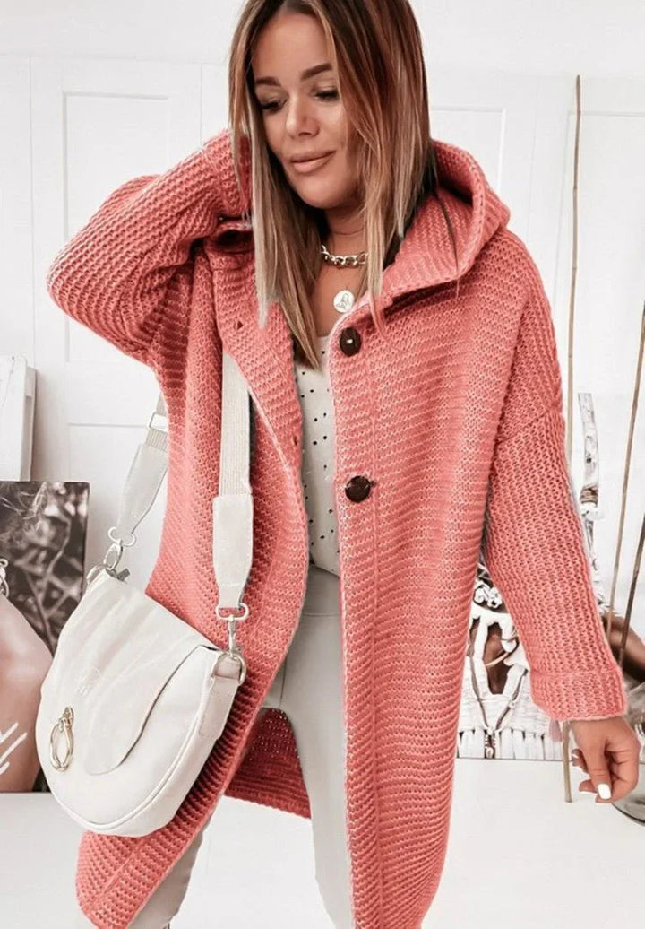 Une femme portant un long cardigan rouge en grosse maille à capuche et gros boutons, assorti à un sac blanc et un pantalon clair, pose dans une pièce lumineuse ornée de portraits artistiques.