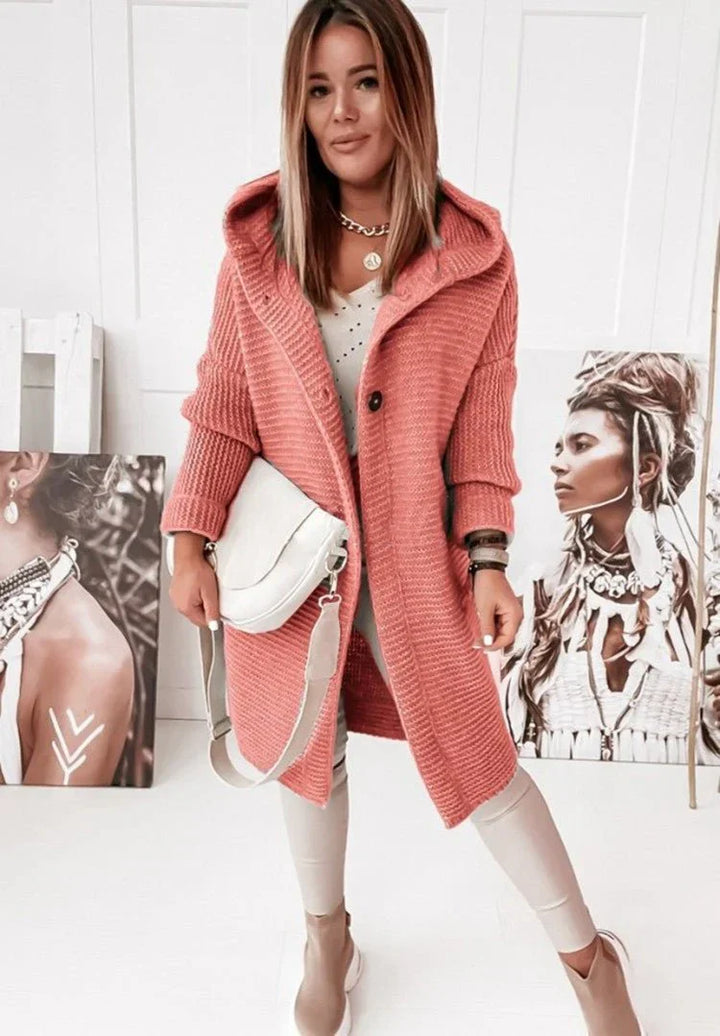 Femme portant un long cardigan rouge saumon en grosse maille, à capuche et gros boutons, assorti à un sac blanc, un pantalon clair et des bottines, posant dans une pièce décorée de portraits artistiques.