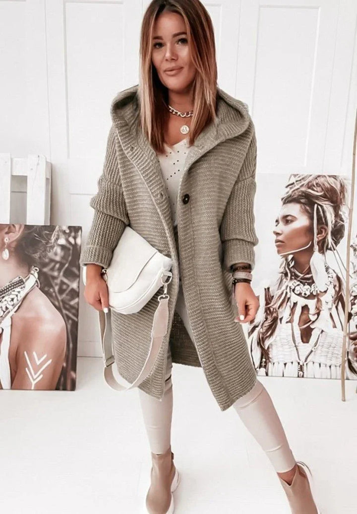 Une femme portant un long cardigan kaki en grosse maille, à capuche et à gros boutons, assorti à un sac blanc, un pantalon clair et des bottines, pose dans une pièce lumineuse décorée de portraits artistiques.