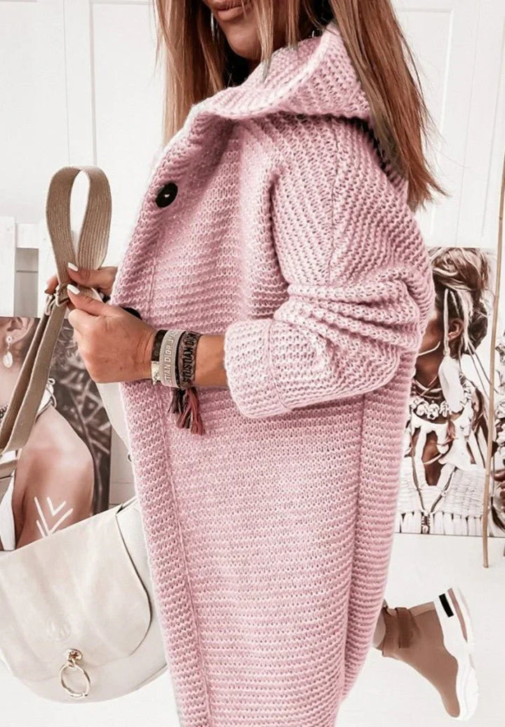 Femme portant un long cardigan en maille épaisse rose avec capuche et manches retroussées, tenant un sac beige et posant dans une pièce lumineuse décorée de portraits artistiques.
