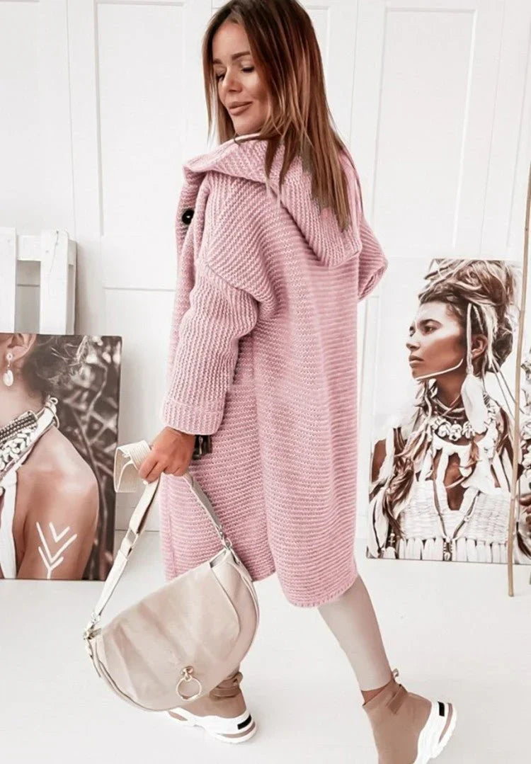 Femme vue de dos portant un long cardigan en maille épaisse rose avec capuche, assorti à un sac beige, un pantalon clair et des chaussures sport, posant dans une pièce lumineuse décorée de portraits artistiques.