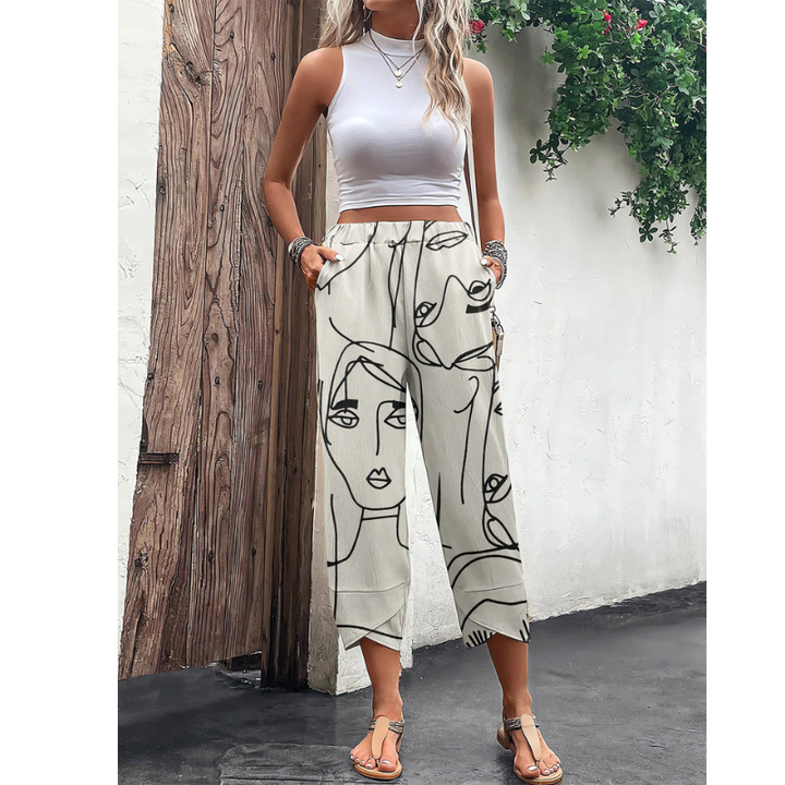 Pantalon ample d’été pour femme de couleur beige clair, avec imprimé artistique de visages en lignes noires, taille élastique avec lien de serrage, coupe fluide longueur cheville, porté avec un top blanc et des sandales en extérieur.