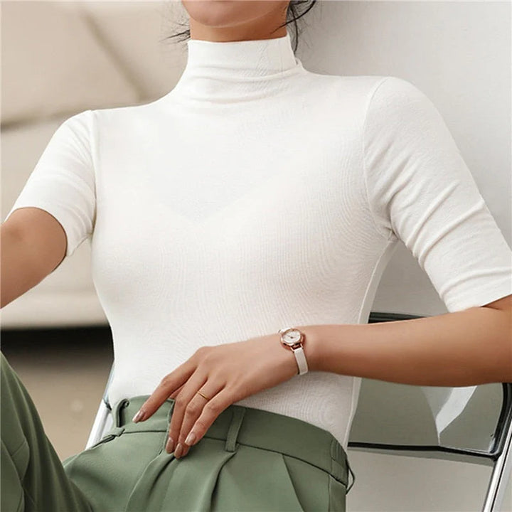 Une femme porte un haut à col roulé ajusté de couleur blanc cassé, à manches courtes. Le tissu lisse et extensible souligne une allure élégante et minimaliste. Le haut est rentré dans un pantalon vert olive à taille haute, créant un contraste raffiné et moderne. Cette tenue dégage un style chic et polyvalent, parfait pour le bureau ou les occasions décontractées.