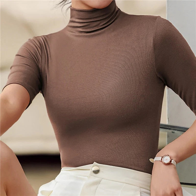 Une femme porte un haut à col roulé ajusté de couleur marron chocolat, à manches courtes. Le tissu lisse et extensible met en valeur la silhouette tout en offrant un confort élégant. Le haut est rentré dans une jupe ou un pantalon beige clair à taille haute, créant une tenue raffinée et moderne, idéale pour le bureau ou les occasions décontractées chic.