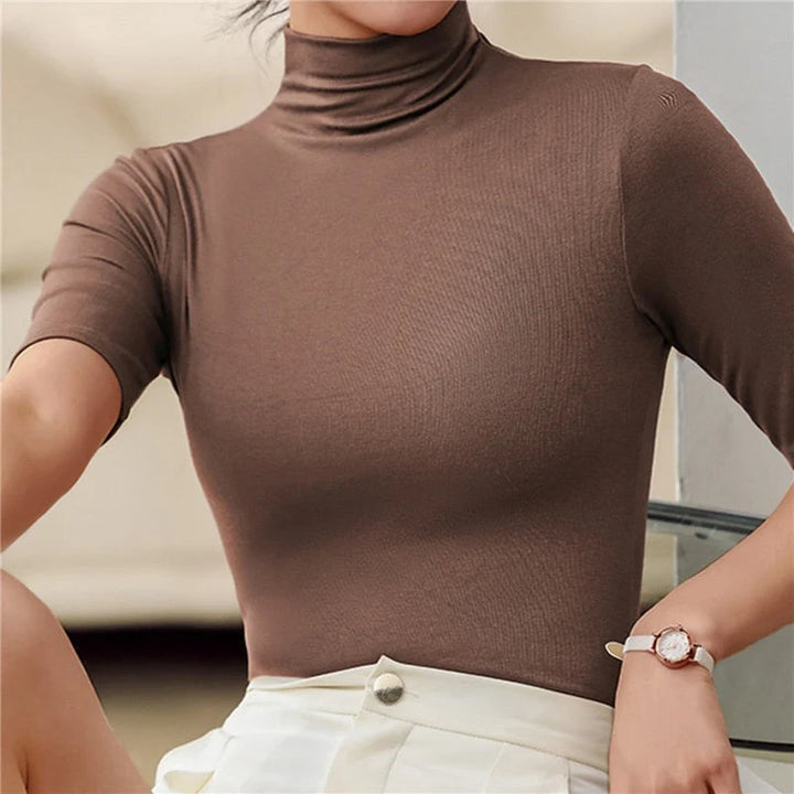 Une femme porte un haut à col roulé ajusté de couleur marron chocolat, à manches courtes. Le tissu lisse et extensible met en valeur la silhouette tout en offrant un confort élégant. Le haut est rentré dans une jupe ou un pantalon beige clair à taille haute, créant une tenue raffinée et moderne, idéale pour le bureau ou les occasions décontractées chic.