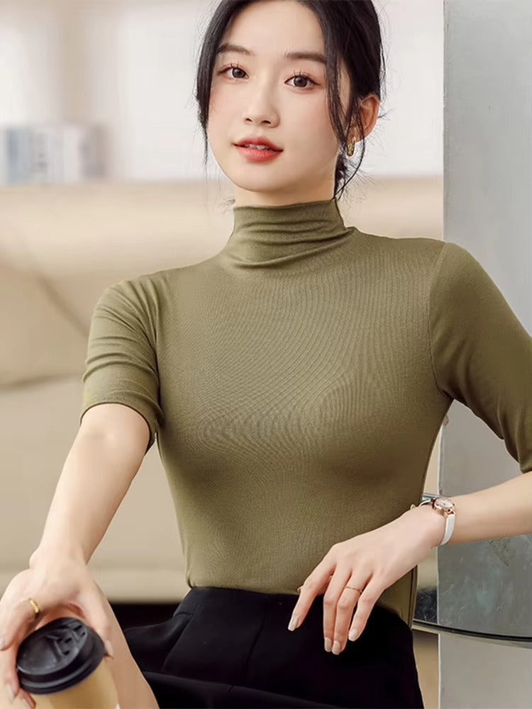 Une femme porte un pull vert à col roulé, ajusté et à manches courtes. Le tissu fin et extensible épouse délicatement sa silhouette, créant une allure élégante et moderne. Assise, une tasse de café à la main, elle porte un pantalon noir taille haute, composant ainsi une tenue chic et minimaliste, idéale pour le bureau ou une sortie décontractée.