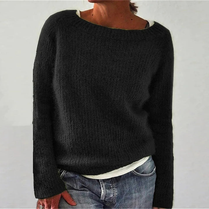 Femme portant un pull en maille noir à coupe décontractée, manches longues et encolure large, assorti d’un t-shirt clair en dessous et d’un jean, photographiée sur un fond neutre.