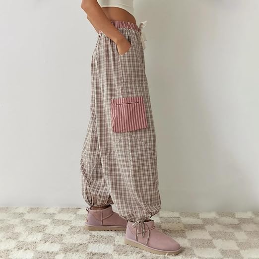 Pantalon de pyjama pour femme vu de profil, à coupe ample, en tissu à carreaux beige et rose, avec taille élastique, poche latérale contrastante et bas resserrés par liens, porté avec des bottines pour un style cosy et tendance.