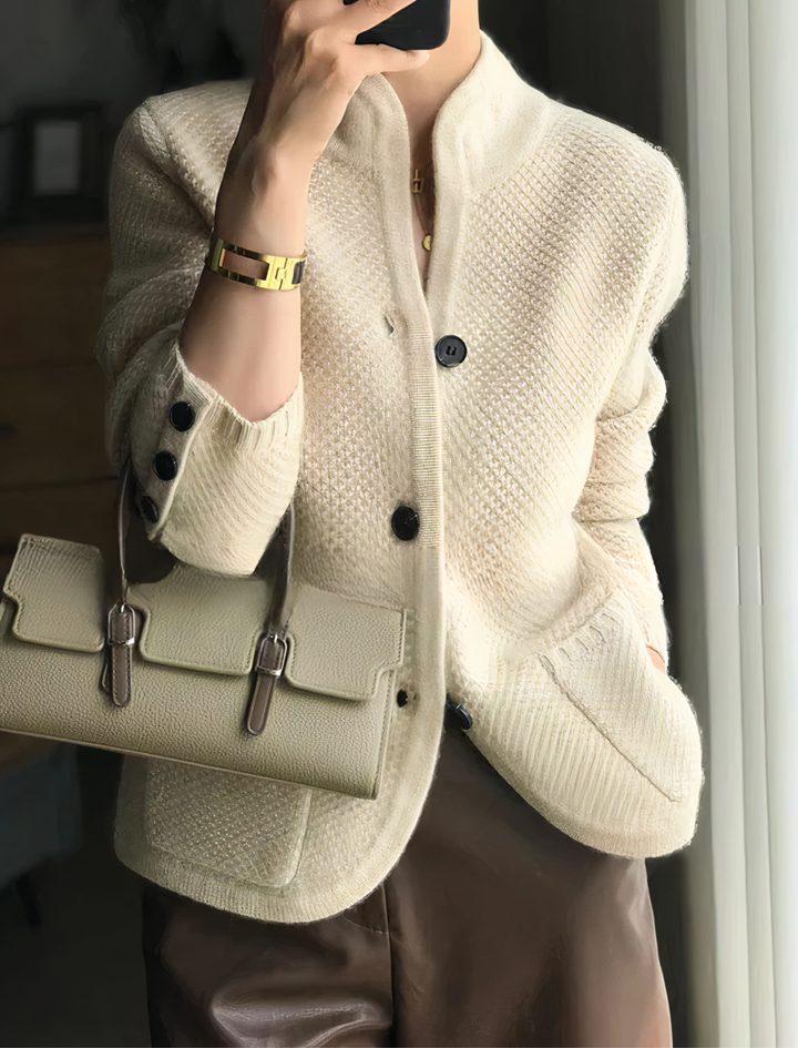 Femme portant un cardigan beige à boutons noirs, avec une coupe moderne, des poches latérales et un col montant, associé à une petite sacoche et un pantalon en cuir pour un look élégant et raffiné.