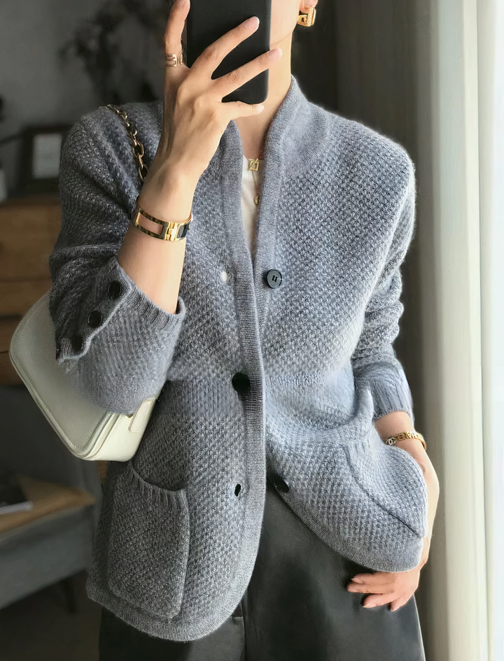 Femme portant un cardigan texturé gris doux, muni de boutons noirs, d’un col montant moderne et de grandes poches avant, assorti à un sac épaule blanc et un pantalon foncé pour un look élégant et contemporain.