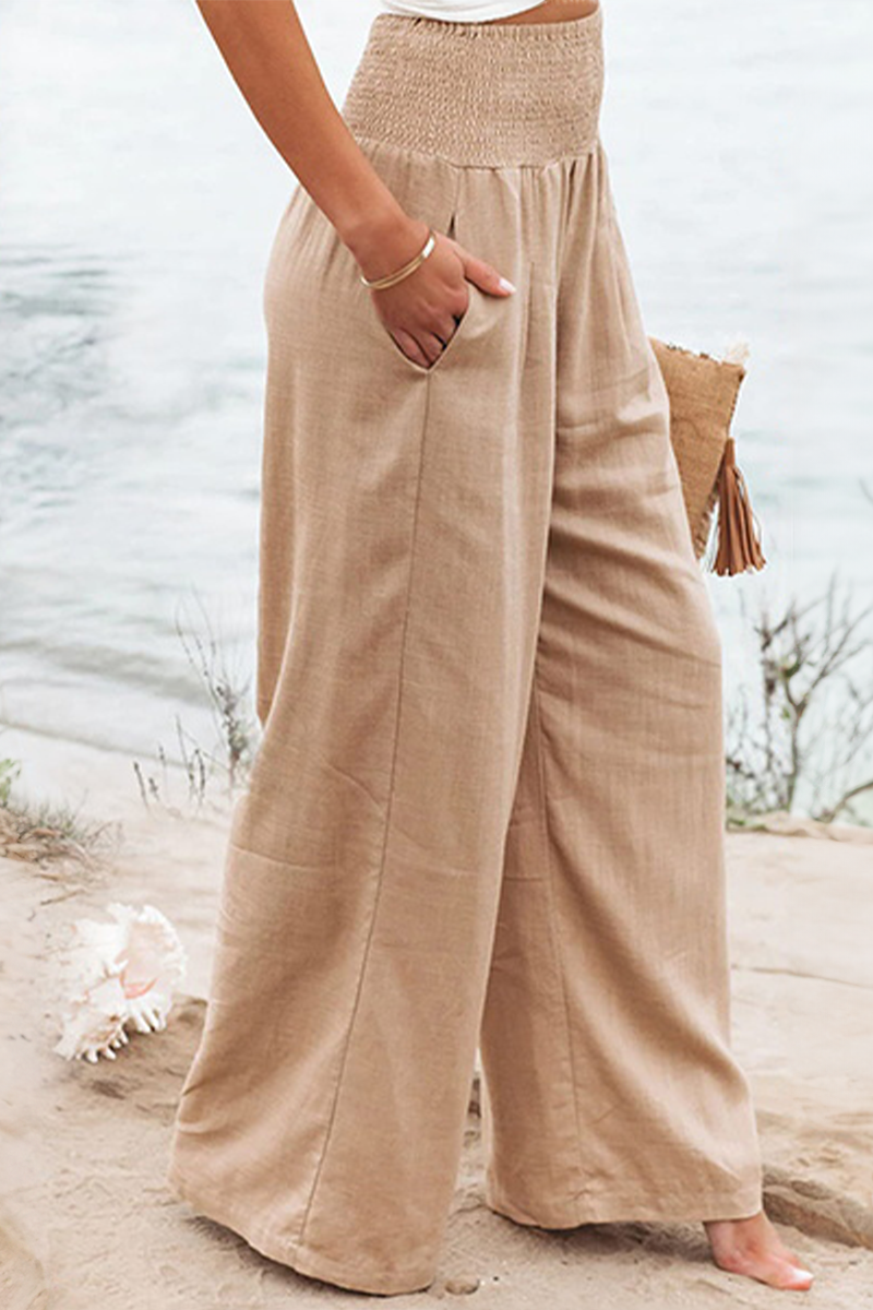Pantalon palazzo pour femme couleur beige sable, taille haute avec ceinture smockée, coupe très large et fluide en tissu léger, porté pieds nus au bord de la mer pour un style estival, naturel et élégant.