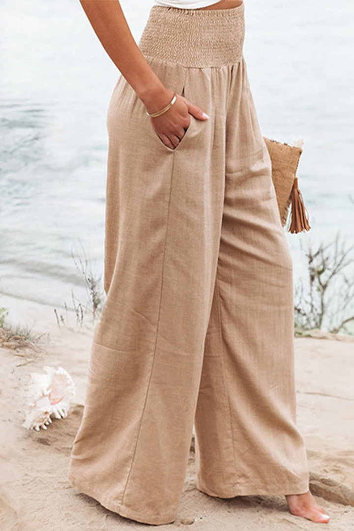 Pantalon palazzo pour femme couleur beige sable, taille haute avec ceinture smockée, coupe très large et fluide en tissu léger, porté pieds nus au bord de la mer pour un style estival, naturel et élégant.