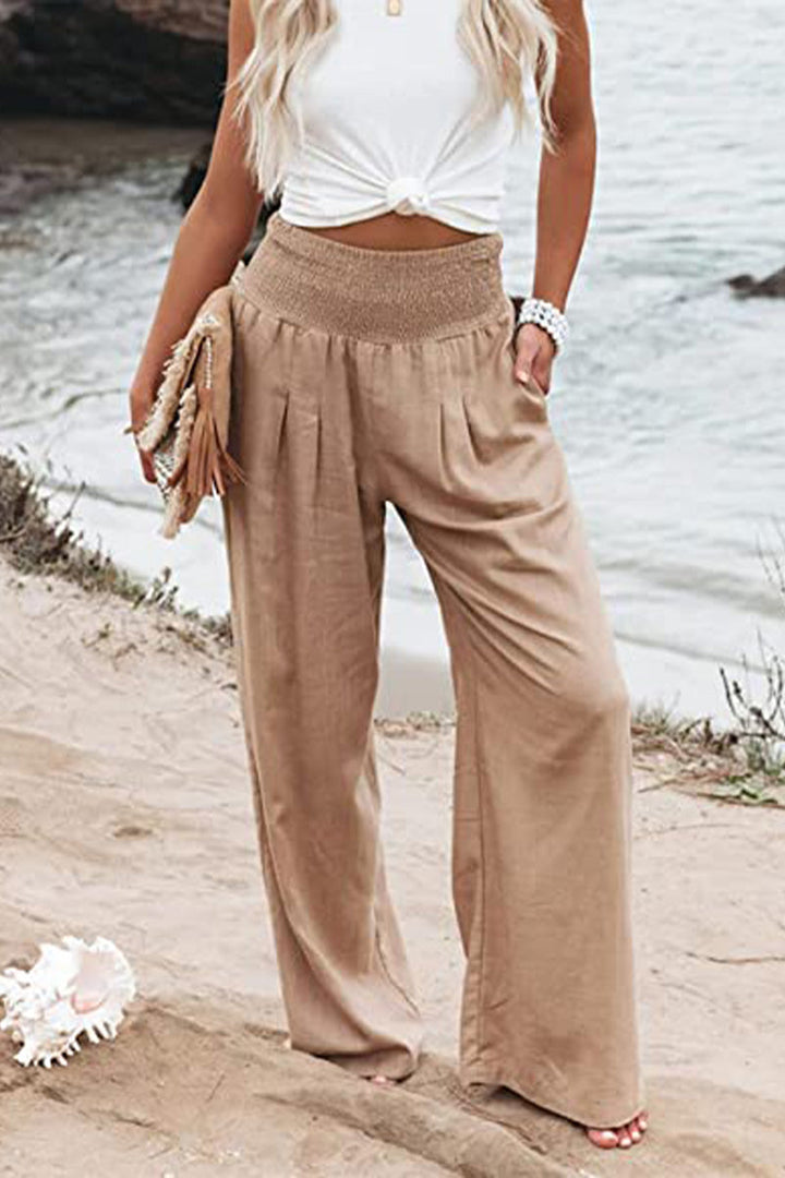 Pantalon palazzo pour femme couleur beige sable, taille haute avec ceinture smockée, coupe très large et fluide en tissu léger, porté au bord de la mer avec un haut blanc noué et une pochette pour un look estival, chic et naturel.