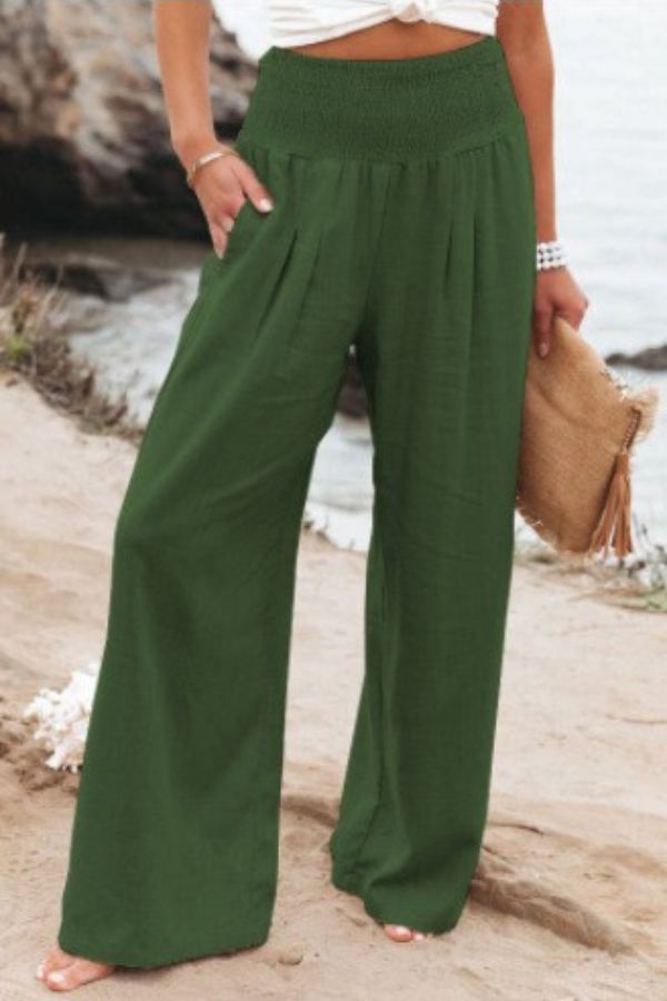 Pantalon palazzo pour femme vert foncé, taille haute avec ceinture smockée, coupe très large et fluide en tissu léger, porté au bord de la mer avec un haut clair pour un style naturel, élégant et estival.