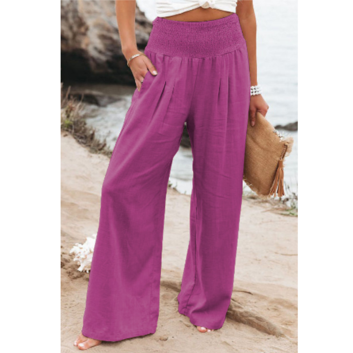 Pantalon palazzo pour femme violet, taille haute avec ceinture smockée, coupe très large et fluide en tissu léger, porté au bord de la mer avec un haut clair pour un style estival, élégant et tendance.