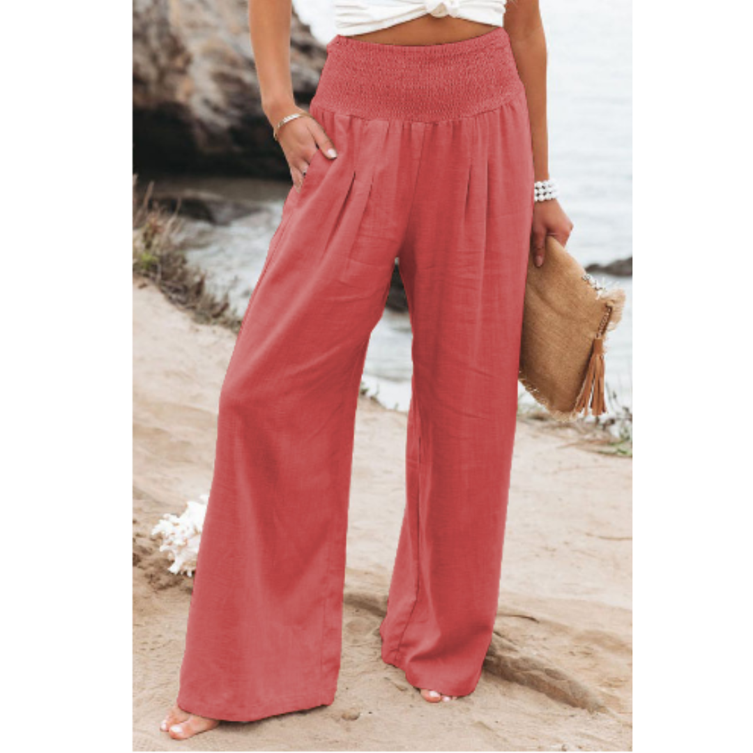 Pantalon palazzo pour femme couleur terracotta, taille haute avec ceinture smockée, coupe très large et fluide en tissu léger, porté au bord de la mer avec un haut clair pour un look estival, chaleureux et élégant.