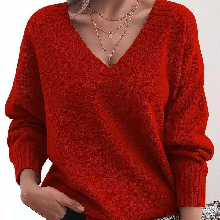 Femme portant un pull en maille rouge à col en V profond avec bords côtelés. Les manches longues sont légèrement amples, et elle porte plusieurs colliers fins en or. Le look est moderne et élégant, avec une touche de simplicité et de féminité.