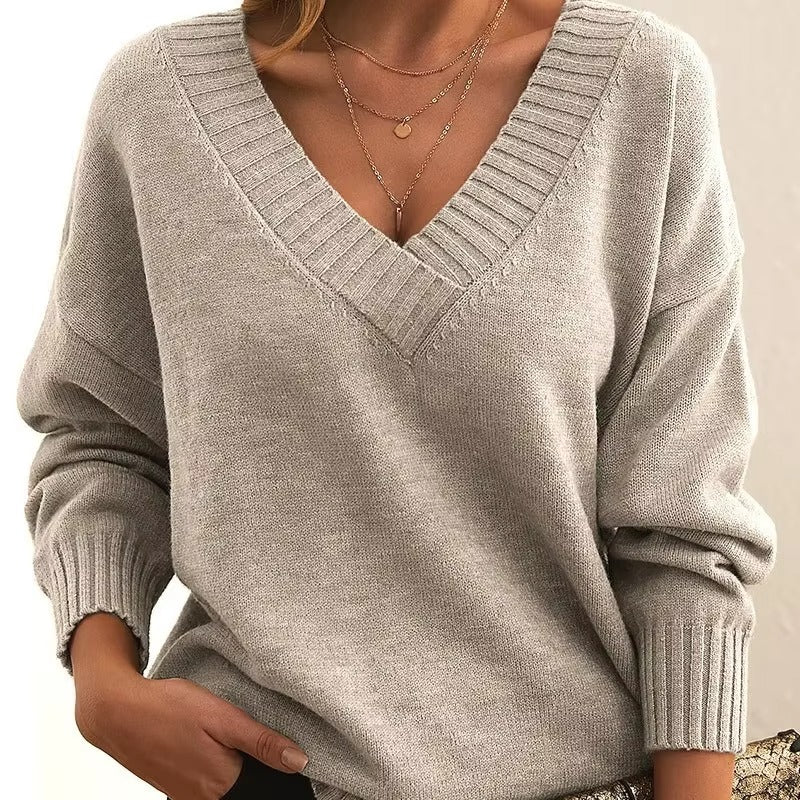 Une femme porte un pull beige à col V en maille côtelée. Les manches sont longues et légèrement amples, et elle arbore plusieurs colliers en or délicats. Son allure est à la fois moderne et élégante, confortable et raffinée, avec une touche de féminité.