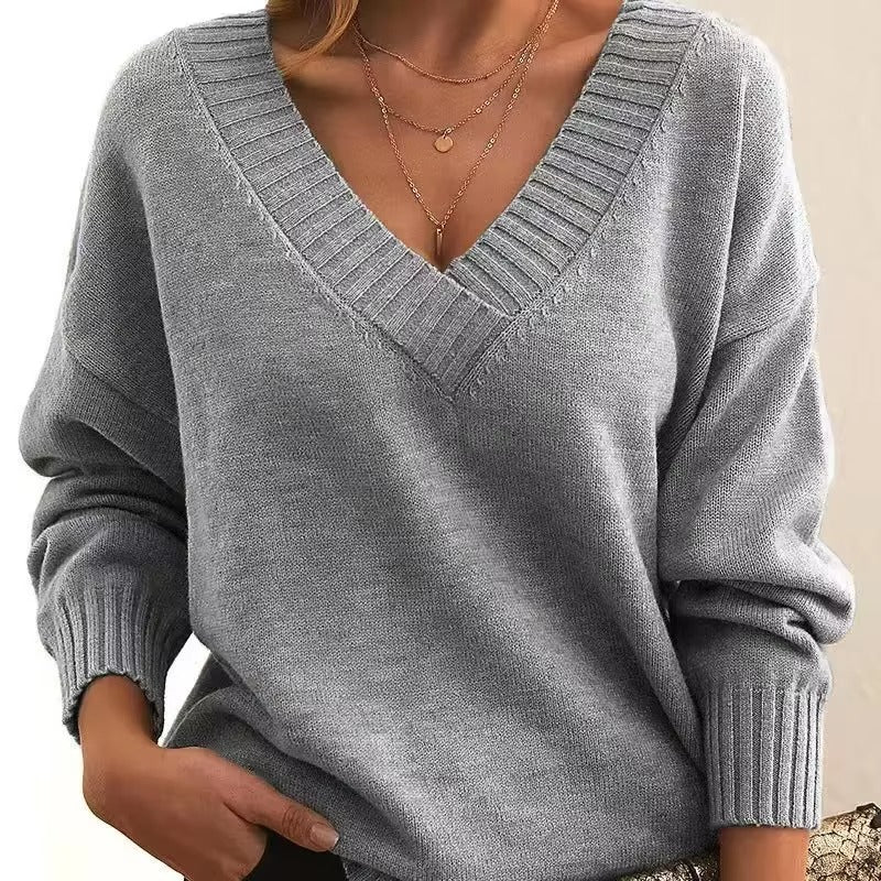 Femme portant un pull en maille gris clair à col en V avec bords côtelés. Les manches longues sont légèrement amples, et elle porte plusieurs colliers fins en or. Ce look moderne et élégant est à la fois confortable et raffiné, avec une touche de féminité.