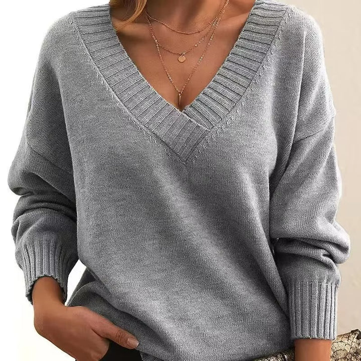 Femme portant un pull en maille gris clair à col en V avec bords côtelés. Les manches longues sont légèrement amples, et elle porte plusieurs colliers fins en or. Ce look moderne et élégant est à la fois confortable et raffiné, avec une touche de féminité.