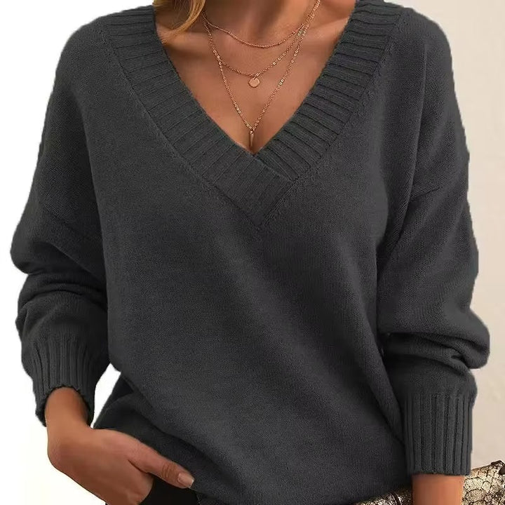 Femme portant un pull en maille gris foncé à col en V avec bords côtelés. Les manches longues sont légèrement amples, et elle porte plusieurs colliers fins en or. Ce pull offre un look moderne et élégant, à la fois confortable et raffiné, avec une touche de féminité.
