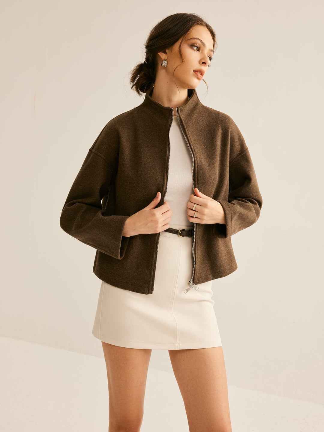 Femme portant un manteau court marron avec col montant et fermeture zippée, associé à une robe courte blanche et une ceinture, créant un look à la fois chic et moderne.