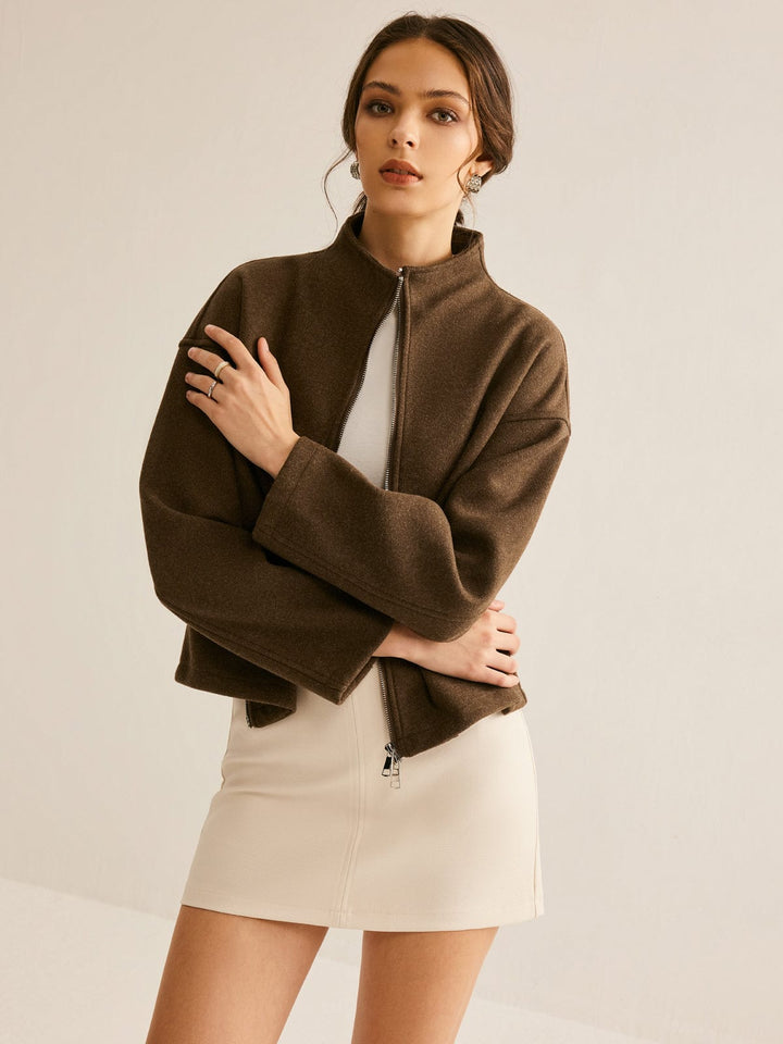 Femme portant un manteau court en laine marron, avec col montant et fermeture zippée, associé à une robe courte beige, pour un look élégant et contemporain.