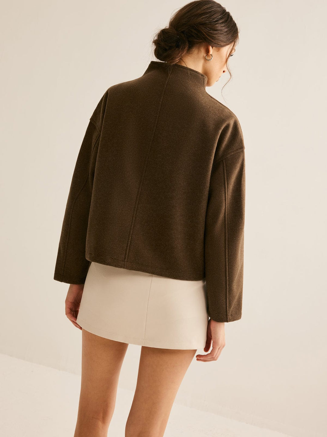 Vue arrière d'une femme portant un manteau court marron à col montant, avec une coupe élégante, associé à une robe beige, pour un look moderne et raffiné.
