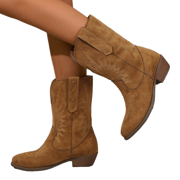 Bottes cowboy en daim marron portées par une femme, présentées en gros plan sur un sol en bois texturé. Le modèle montre des broderies décoratives et un talon bas, créant un style western tendance et décontracté.