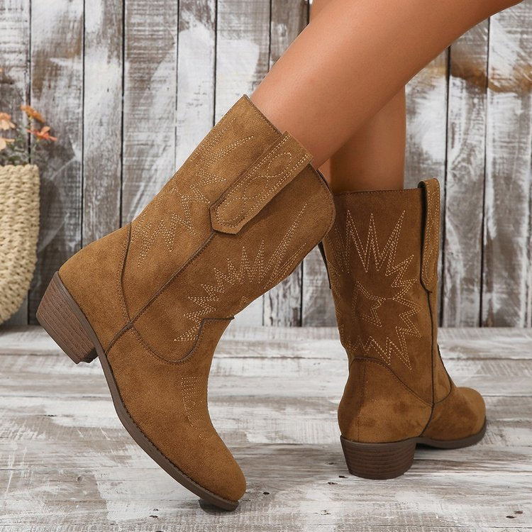 Bottes cowboy en daim marron portées par une femme, mises en valeur dans un décor en bois rustique. Les bottes présentent des broderies détaillées, une tige mi-haute et un talon bas, offrant un style western moderne et tendance.