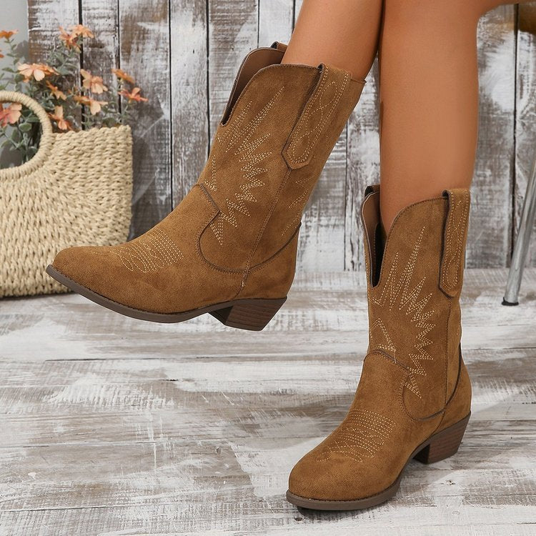 Bottes cowboy en daim marron portées par une femme, mises en valeur avec des broderies décoratives et un petit talon. Les bottes sont présentées dans un décor rustique en bois, avec un style western moderne et tendance.