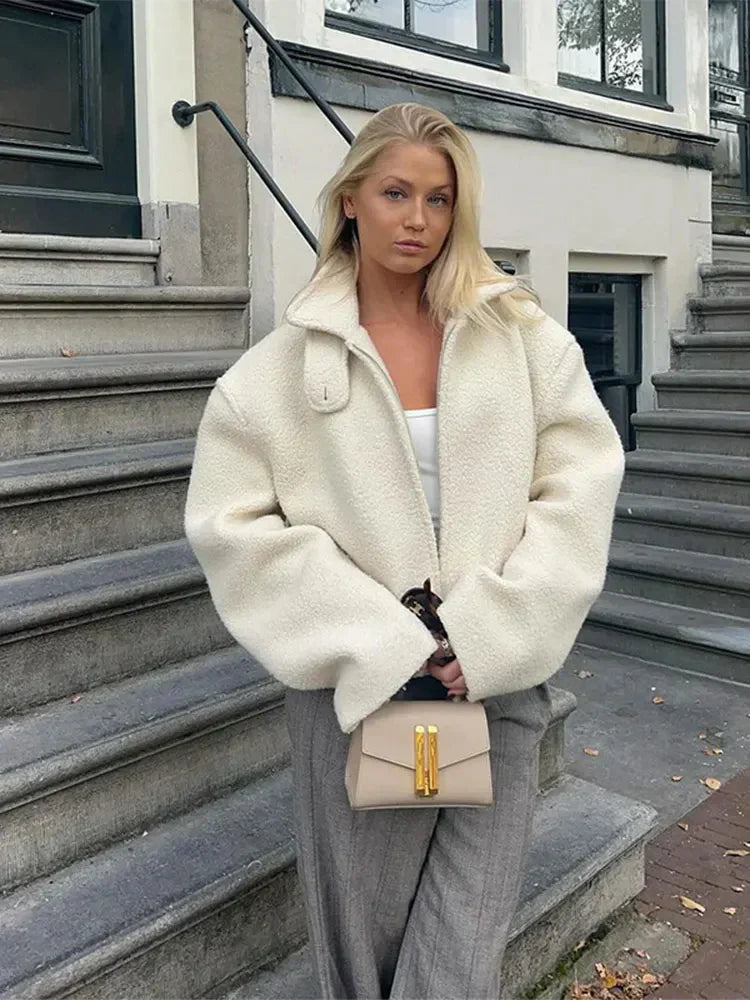 Une femme élégante pose en extérieur sur des marches en pierre, vêtue d'une veste minimaliste en laine épaisse blanche. Elle porte un pantalon gris à jambes droites, un simple haut blanc et un petit sac à main beige à fermoir doré. En arrière-plan, on aperçoit une façade urbaine moderne avec des fenêtres et un escalier.