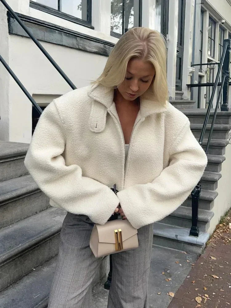 Une femme se tient sur le trottoir, devant un escalier en pierre. Elle porte une veste chaude en laine bouclée blanche, au style épuré et moderne. Son regard est baissé, un petit sac à main beige à fermoir doré à la main. Un pantalon gris droit et un simple haut blanc complètent sa tenue minimaliste, idéale pour l'hiver.