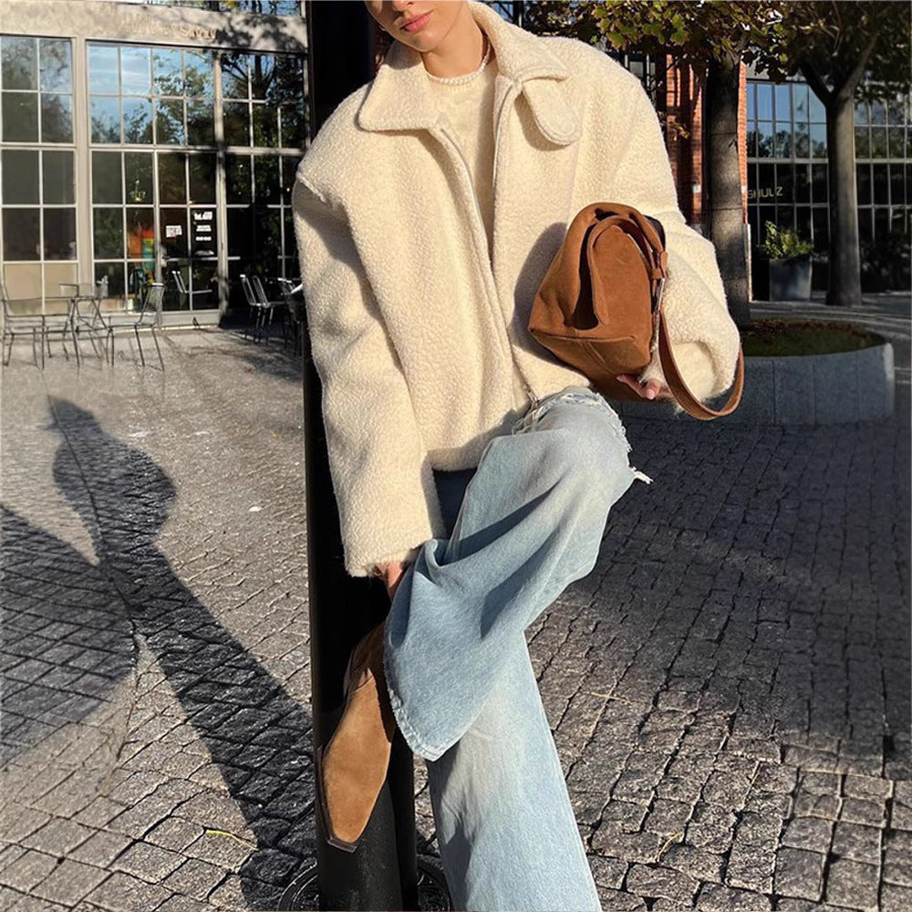 Une femme est assise en terrasse sur une rue pavée, profitant d'une journée ensoleillée. Elle porte une épaisse veste en laine bouclée blanche, assortie à un pull, un jean évasé délavé et des bottines marron. Un sac en daim marron complète sa tenue, créant un look hivernal minimaliste, élégant et confortable dans un cadre urbain moderne.