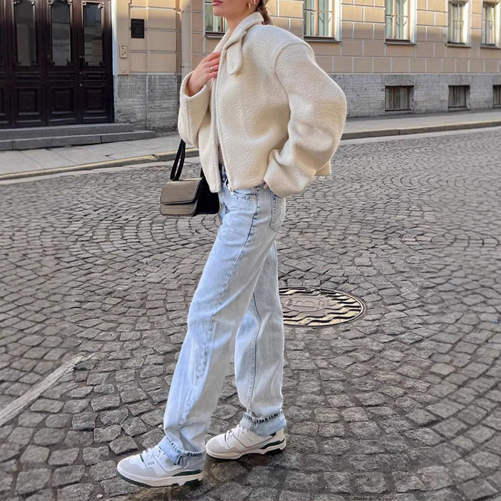 Une femme marche dans une rue pavée, vêtue d'une veste minimaliste en laine bouclée blanche épaisse. Elle porte un jean droit délavé, des baskets blanches et un sac à main beige et noir. Sa tenue dégage une allure à la fois décontractée et élégante, idéale pour l'hiver en ville.
