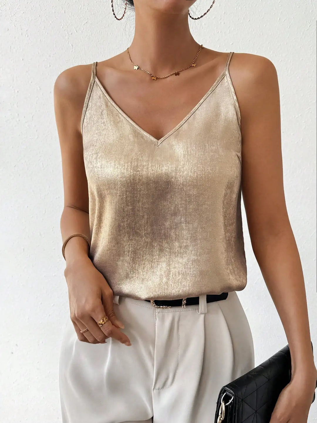 Débardeur élégant en doré, à fines bretelles et col en V, porté avec un pantalon blanc fluide et une ceinture noire. Un look moderne et chic, parfait pour une tenue décontractée et raffinée.